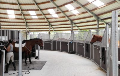 A horse is being tacked up by a rider in the center of a round, covered barn, with stalls on the outside