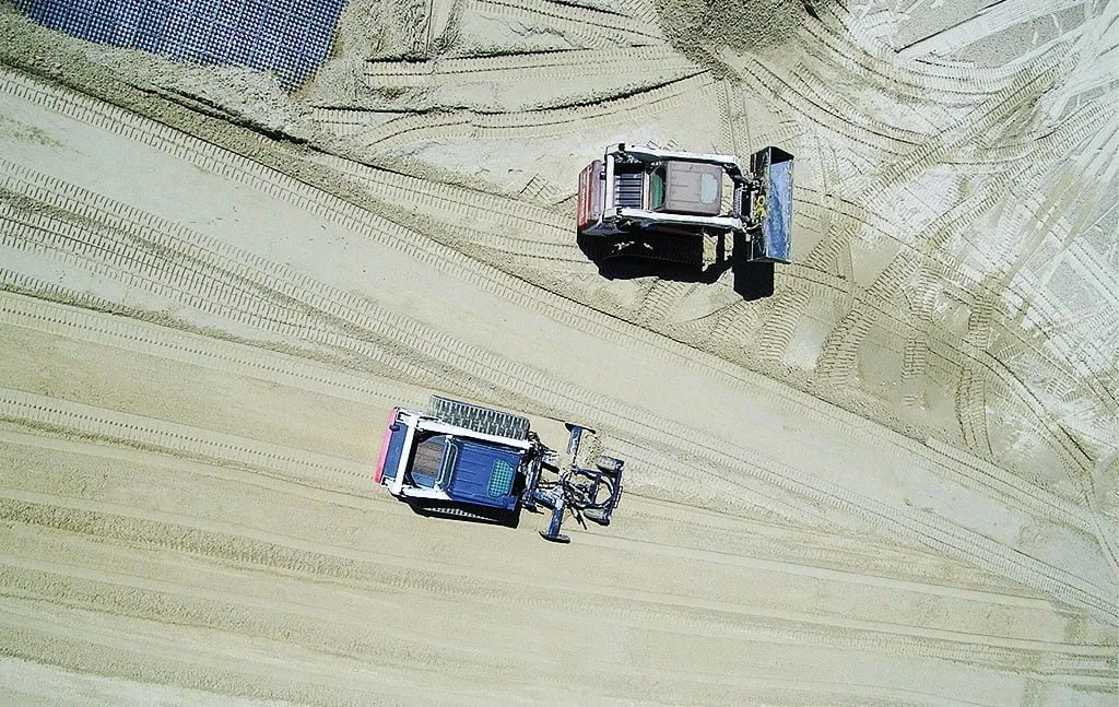 An aerial view of construction equipment spreading and evening sand in an outdoor arena.