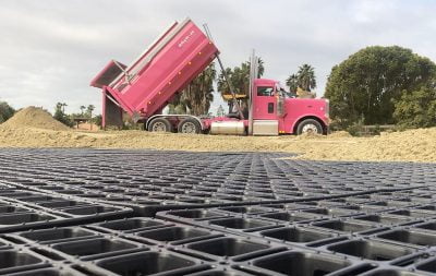 A pink construction truck dumps sand in an outdoor arena that's under cpnstruction.